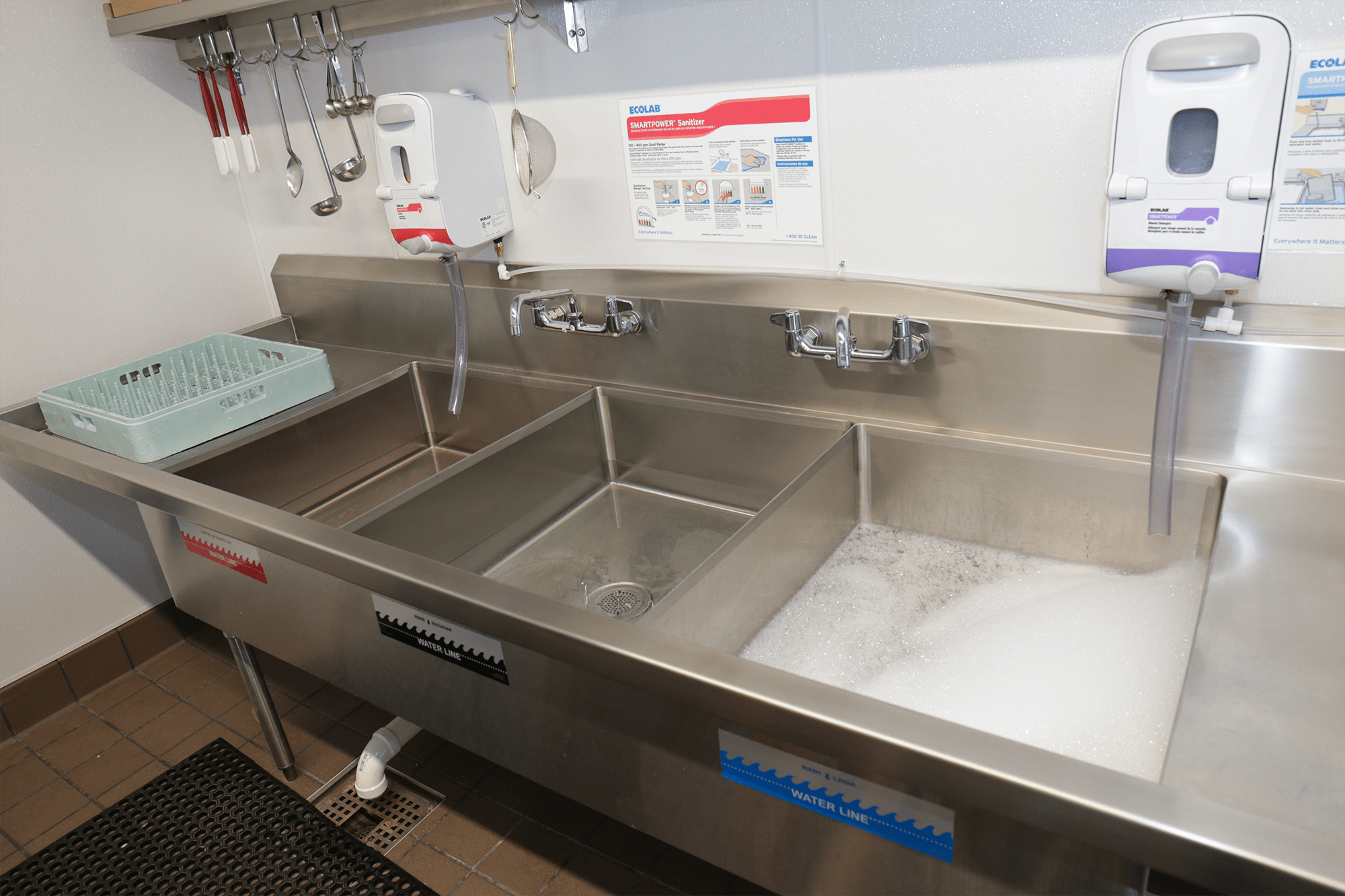 Stainless-steel sink with 3 compartments. From right to left, foamy detergent water, clear rinse water, and pink sanitizer water.  The end has a light blue drying rack. 