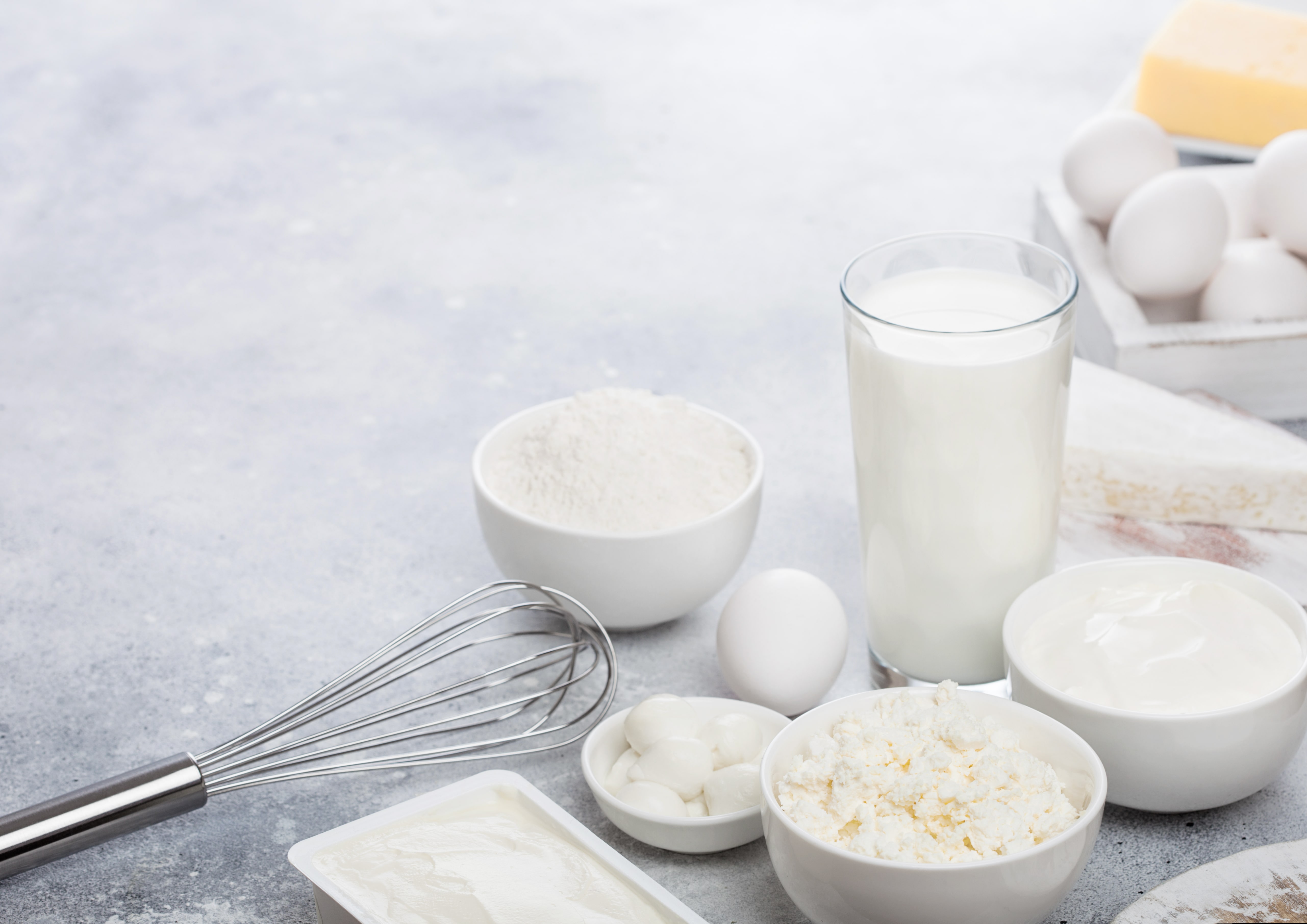 Fresh dairy products on white table background. Glass of milk, bowl of sour cream, cottage cheese and baking flour and mozzarella. Eggs and cheese. Steel whisk